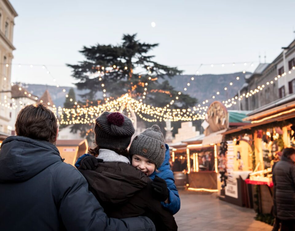 Weihnachtsmarkt Meran mit beleuchteten Ständen und Blick auf die verschneiten Bergspitzen